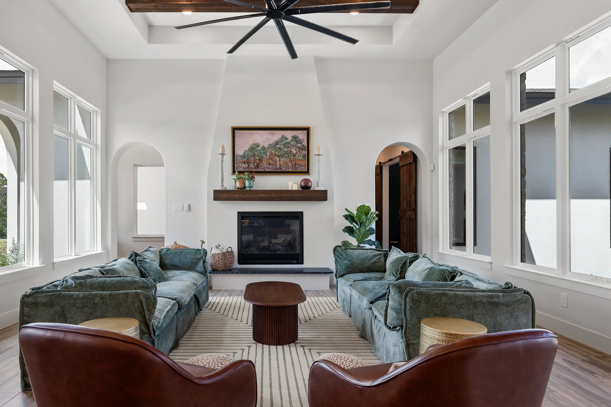 Bright living room with large windows, wood ceiling beams, cozy green sofas, and a modern fireplace in a custom home located in Mitchell Ranch, Bulverde Texas.