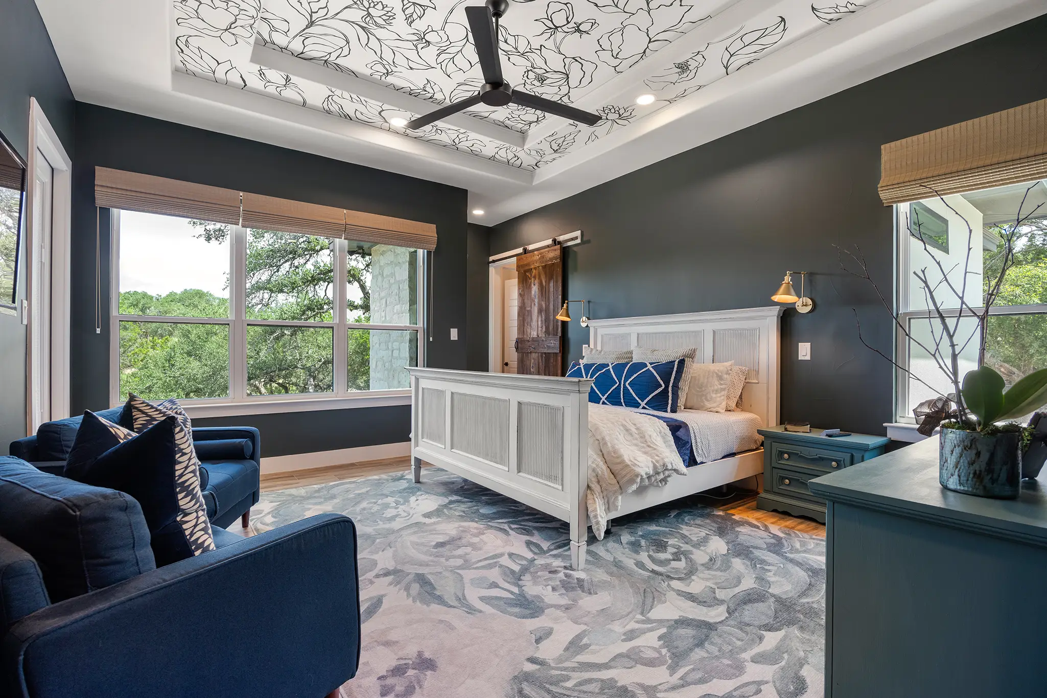 Primary bedroom with dark accent walls, patterned tray ceiling, large windows, and a light wood bed frame inside a custom home in Mitchell Ranch, Bulverde Texas.