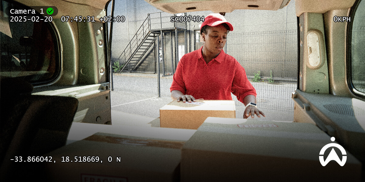 A delivery person wearing a red uniform and cap stands outside a van, preparing to handle cardboard boxes in a parking area.