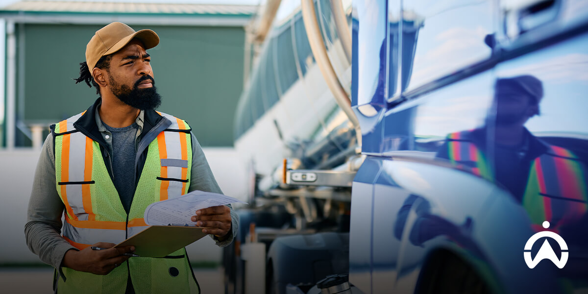 A worker in a high-visibility vest holds a clipboard, conducting vehicle maintenance checks on a shiny blue vehicle in a well-lit industrial area.
