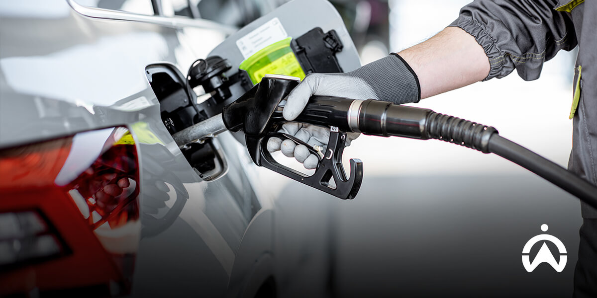 A person in a gray glove fuels a silver car at a gas station, holding the nozzle near the fuel cap.