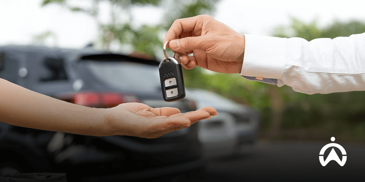A hand transferring car keys to another hand, set against a blurred background of parked vehicles.