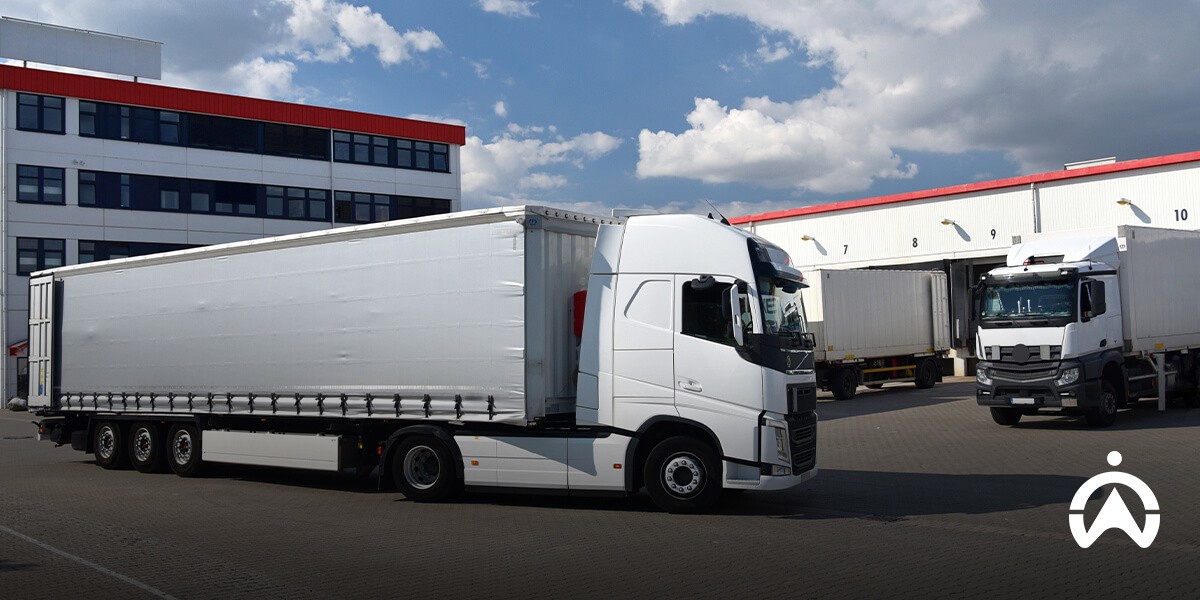 A white truck parked outside a warehouse, with another truck in the background under a partly cloudy sky.