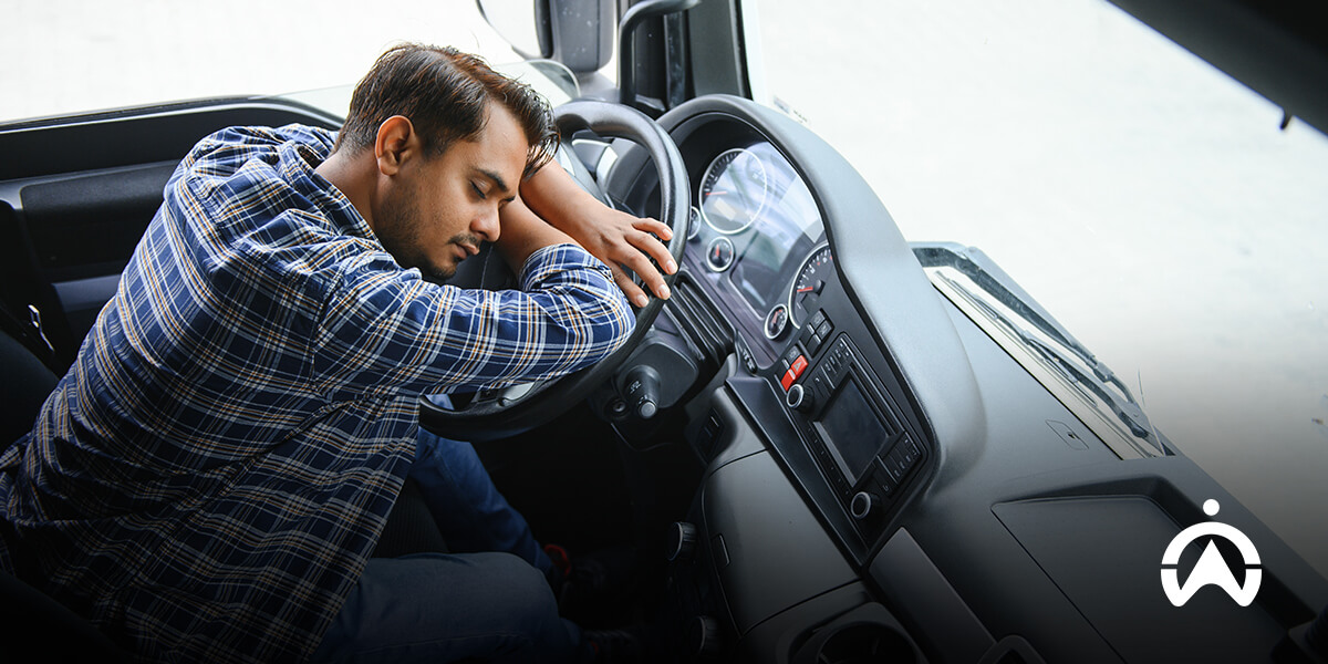 A driver rests his head on the steering wheel of a truck, showing signs of fatigue inside the vehicle's cabin.