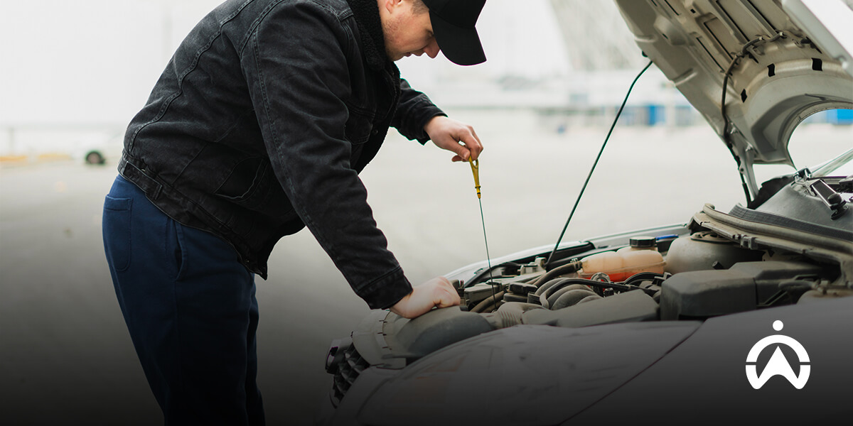 A person in a black jacket and cap inspects the engine of a car with the hood raised, checking the oil level with a dipstick.