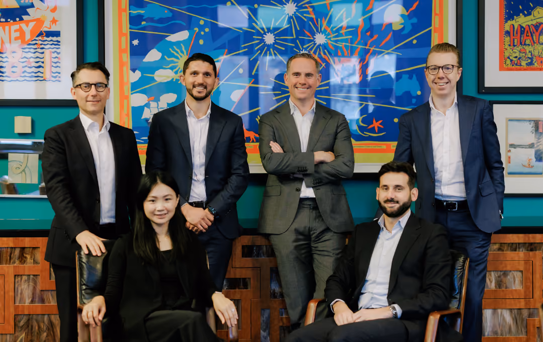 Group of six business professionals, four standing behind and two seated in chairs, in a colorful office.
