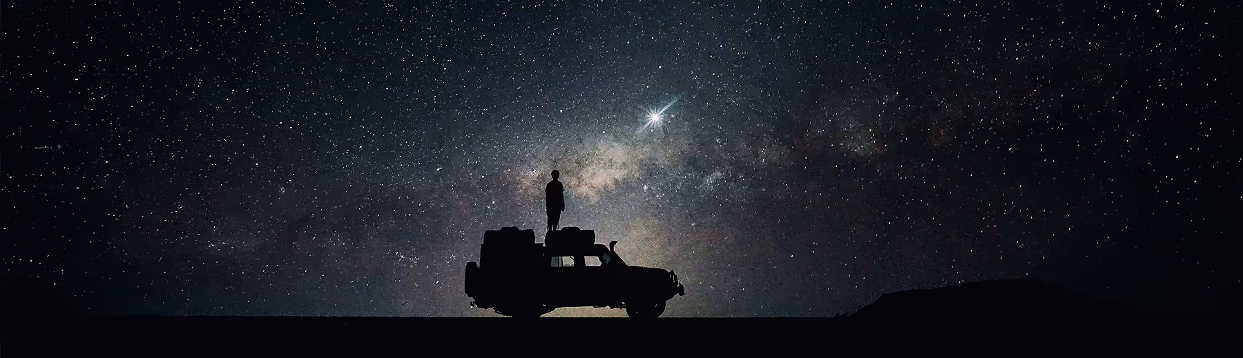 The silhouette of a man standing on top of his off road vehicle beneath an endless starry sky.
