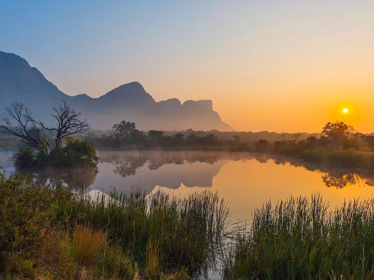 A dam at sunrise surrounded with reeds and the Limpopo landscape and mountains in the distance. 