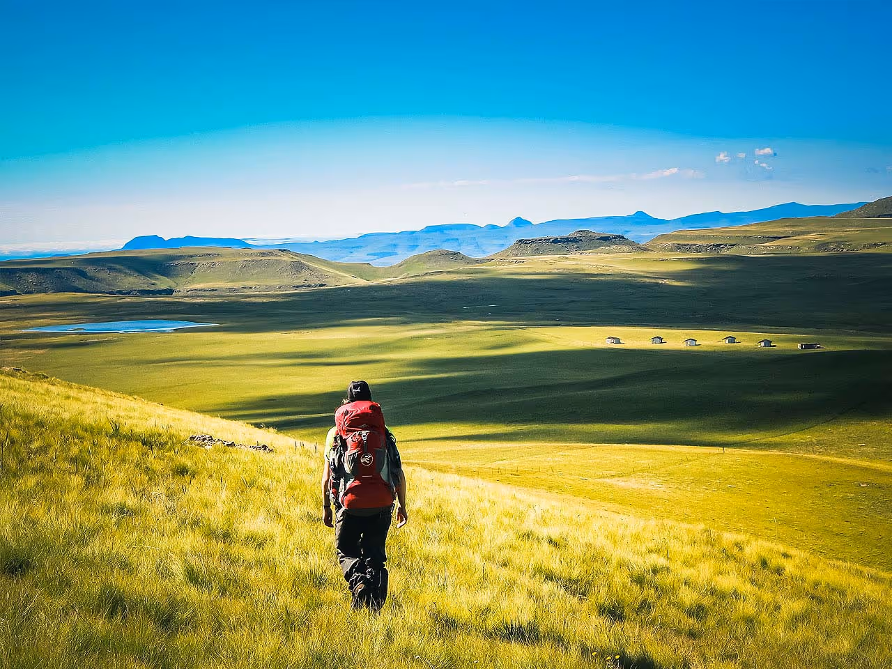A man hiking in a field of grass overlooking some huts, a dam and the Lesotho mountains in the background.