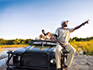 A safari vehicle parked on a sand bank with a guide and tracker pointing out something in the distance to guests.