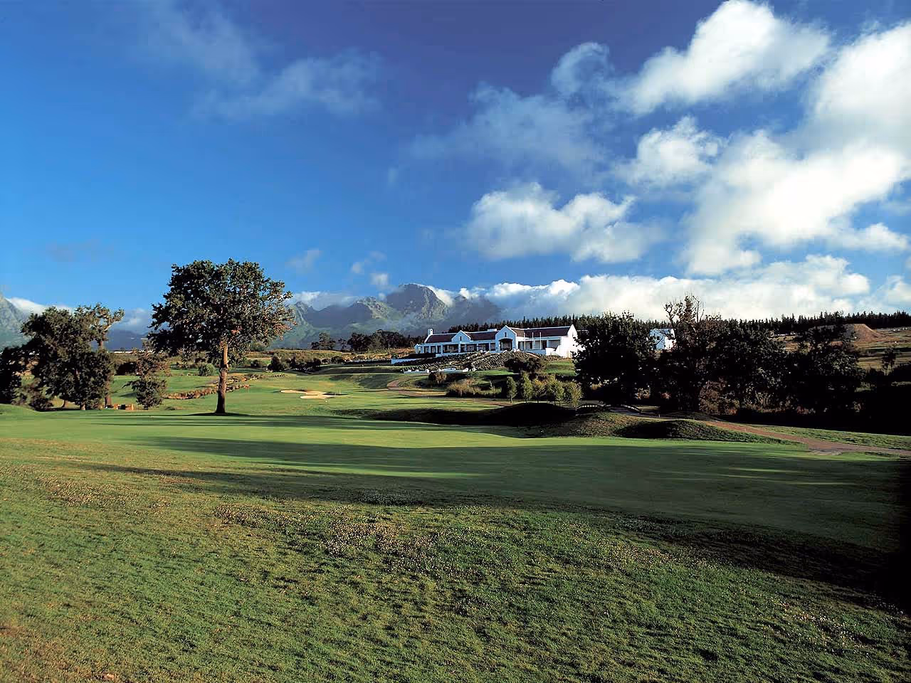 A view of a Cape Dutch manor on a golf course with mountaina in the distance.