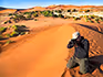 A man with his camera on his knees in the sand on a dune taking a picture with more dunes behind him.