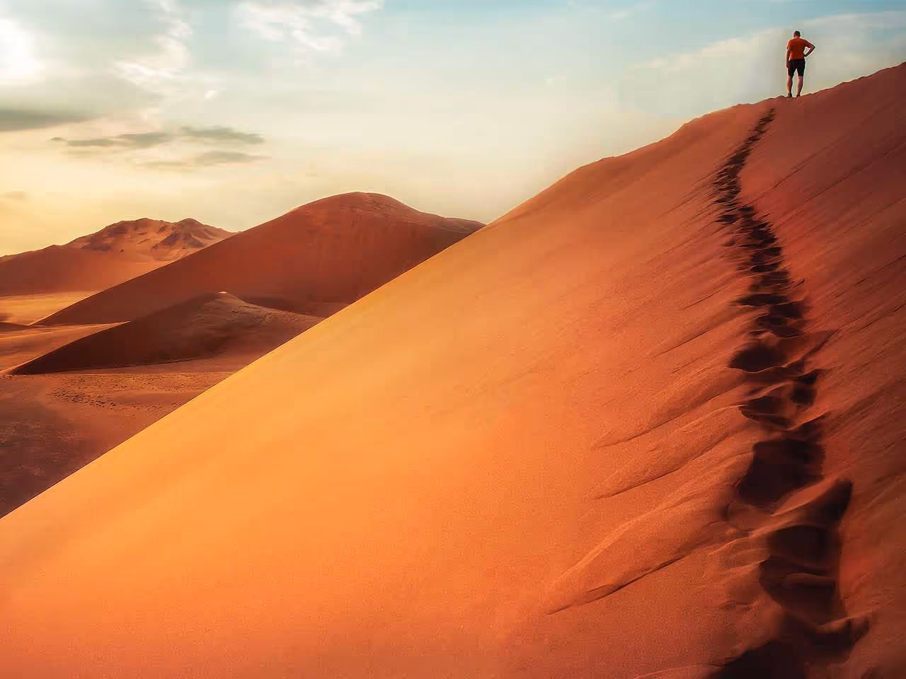 A view from a dune of a man at the top climbing up and more towering sand dunes in the background.