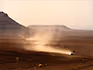 A lone vehicle driving on a long winding gravel road leaving dust in the distance. 