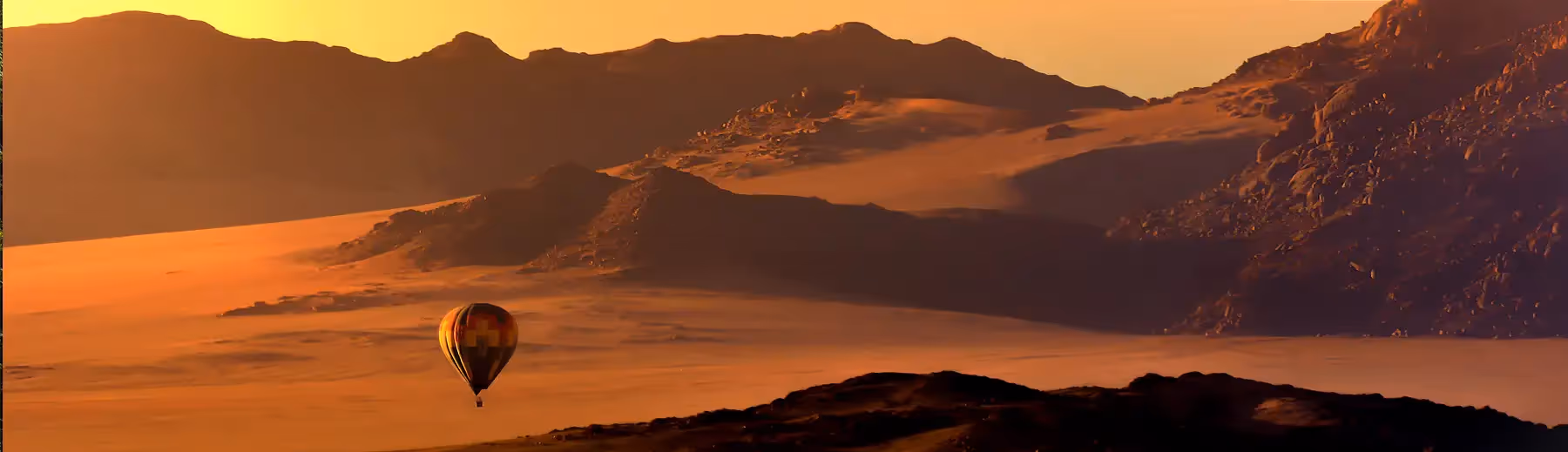 A hot air balloon floating above the desert landscape of the Namib Desert in front of the distant mountains. 