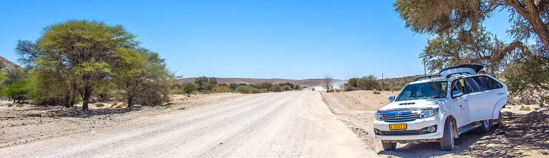 A Namibia Tours & Safaris Fortuner vehicle parked roadside beneath a tree and another vehicle approaching in the distance.