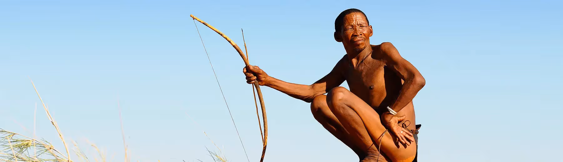 A traditional San bushman on his haunches holding his bow and arrow glaring into the sun.