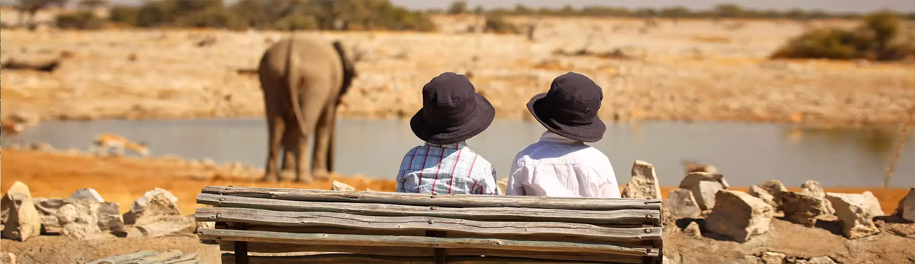 Two children sitting on a wooden bench watching an elephant at a waterhole in Etosha National Park.