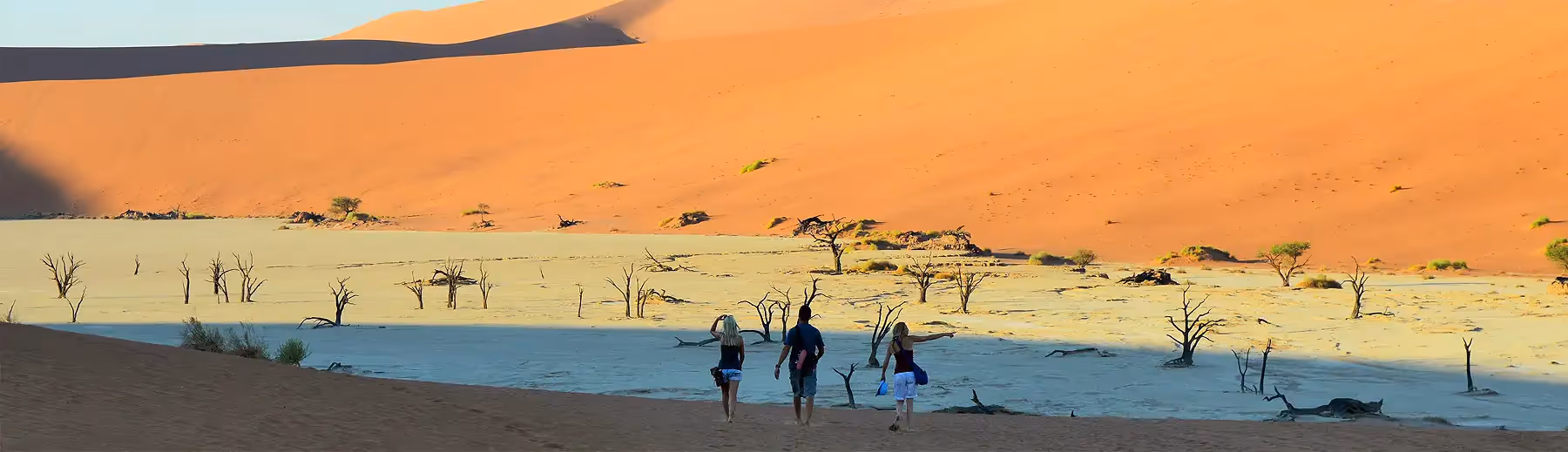 Three people walking down a red sand dune onto the dry pan at Dead Vlei with dead trees and surrounded by more sand dunes. 