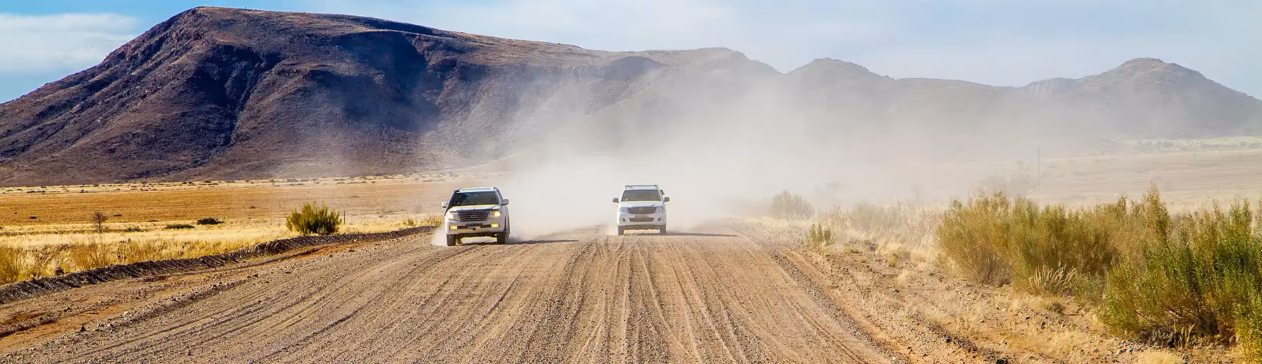 A panoramic view of two 4x4 vehicles driving alongside one another on a gravel road leaving a trail of dust. 