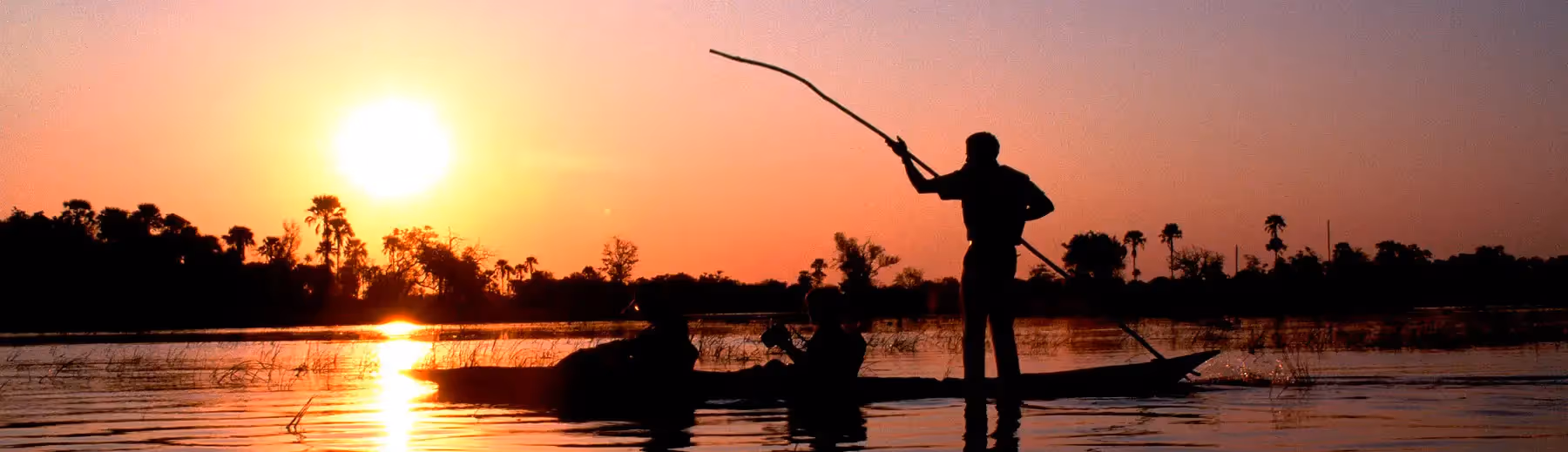 A silhouette of two people being poled along on a mokoro excursion on the river at sunset. 