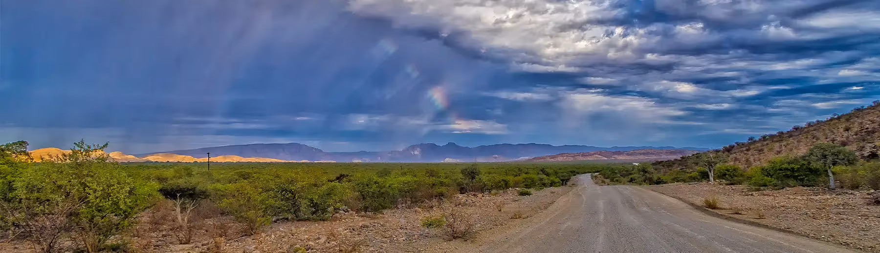 A panoramic view of a gravel road in Damaraland where the sun is shining through the clouds with the hint of a rainbow. 