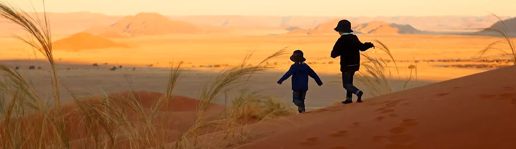 Two children running down a red sand dune with the endless desert landscape in the distance. 