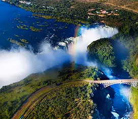 An aerial view over Victoria Falls showing the spray of the falls and the bridge over the gorge with a rainbow.