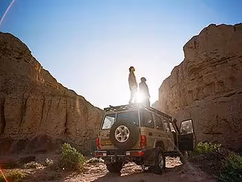 Two people standing on top of a land cruiser looking up at the surrounding sand cliffs. 