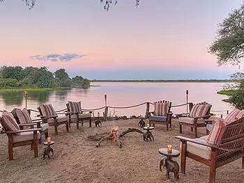 A fire pit surrounded by comfortable seating on a sandy island right on the river at sunset.