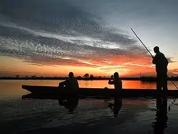 The silhouette of two people on a mokoro being poled along the river at sunset.