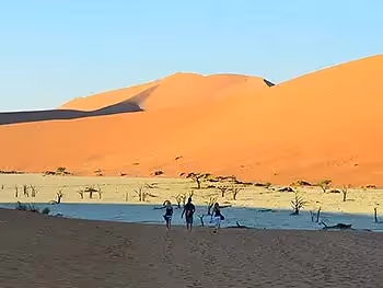Three people walking down a dune into the white salt pan of Dead Vlei with a red sand dune in the background.