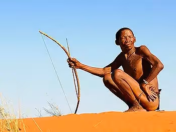 A bushman with bow in hand crouched on a Kalahari dune looking into the sun.