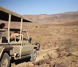 A side view of an open safari vehicle driving on a gravel road in Damaraland with mountains in the background.