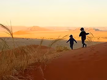 A silhouette of two children running down a red sand dune with the desert landscape in the background. 