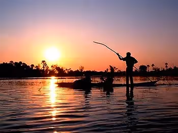 The silhouette of two guests and a poler on a mokoro on the river at sunset.