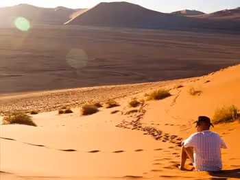 A man sitting on a sand dune looking out over the desert landscape and dunes in the distance. 