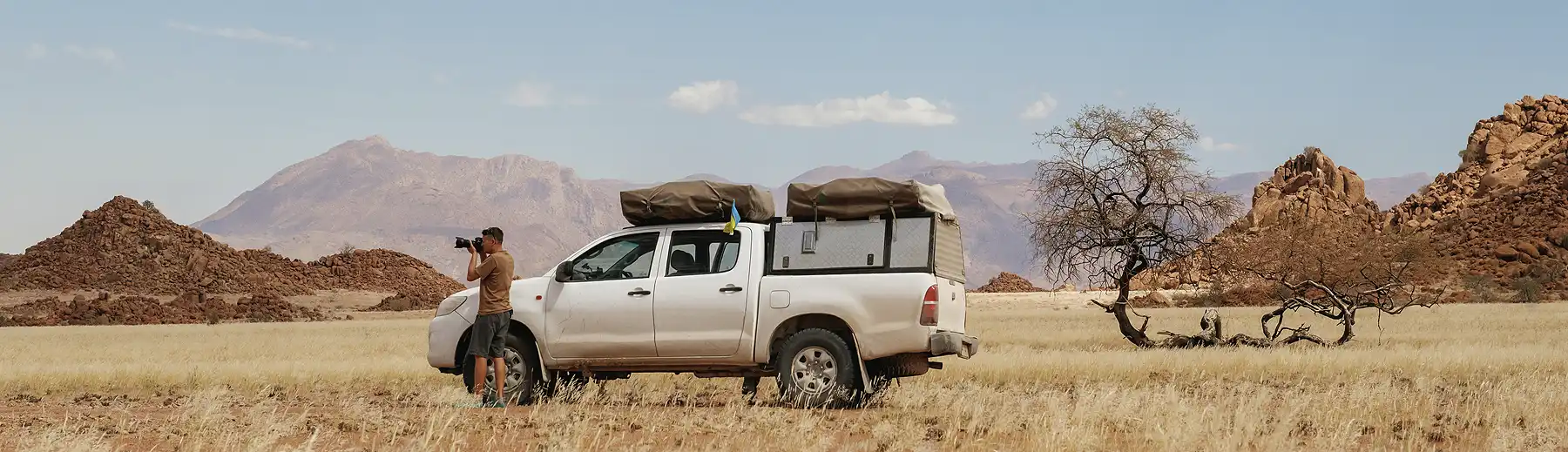 Man photographing desert landscape beside white 4x4 vehicle in Namibia, with rocky hills and a mountain in the background.