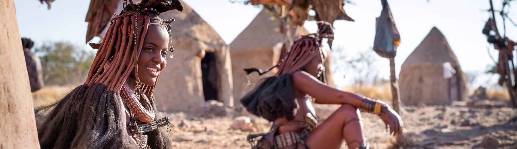 Himba women sitting outside a traditional village in northern Namibia, wearing traditional ochre hairstyles, jewellery and goatskin clothing.