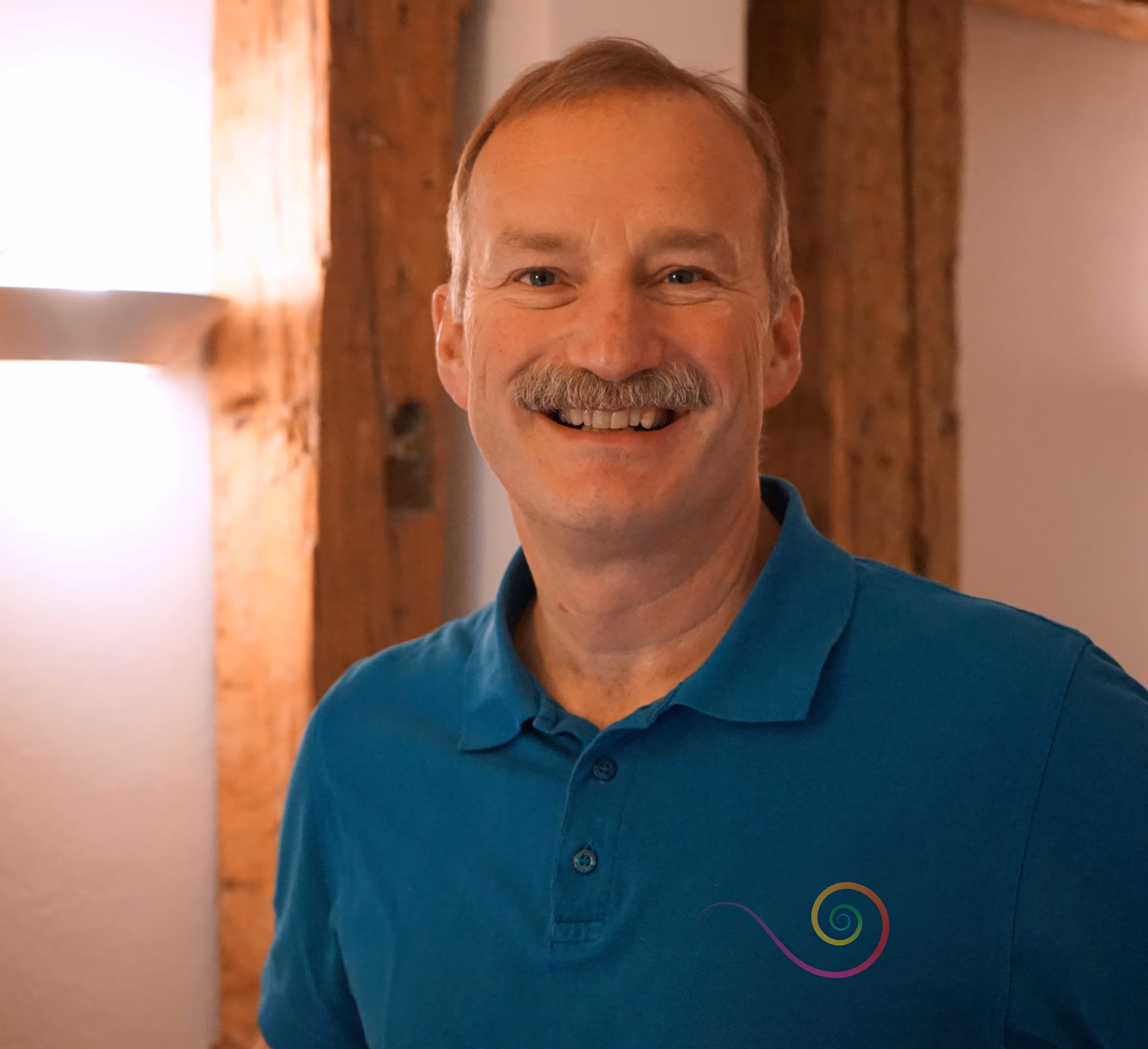 Portrait Dr. med. Thomas Meider-Lenschow, lacht mit Schnauzbart und blaugrünem T-Shirt mit bunter Logo-Praxisschnecke