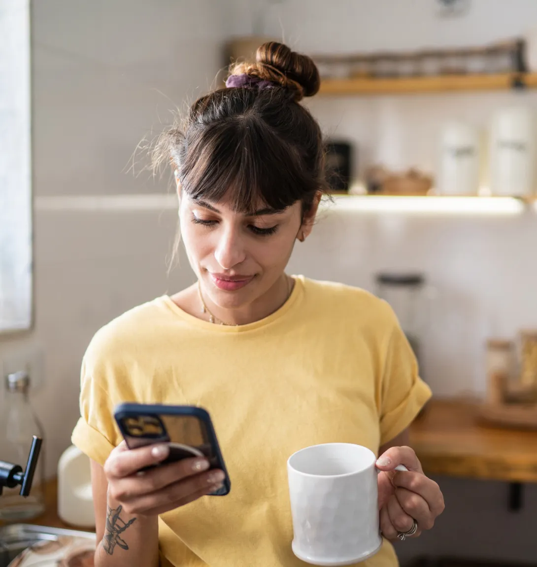 Woman in a yellow t-shirt using a phone while holding a cup in her other hand
