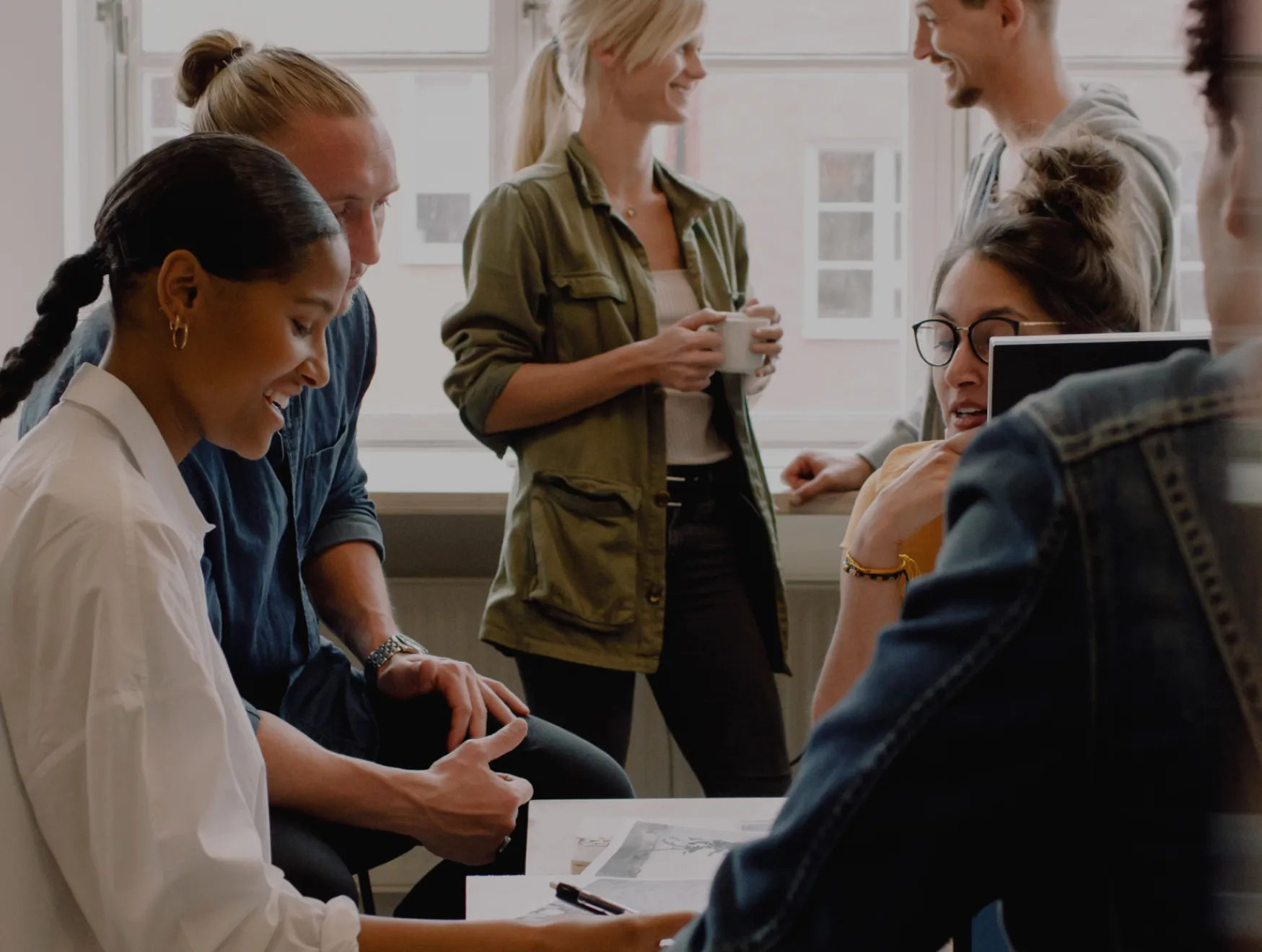 Group of coworkers gathered around a table in a casual meeting, reviewing papers while others stand nearby holding coffee.