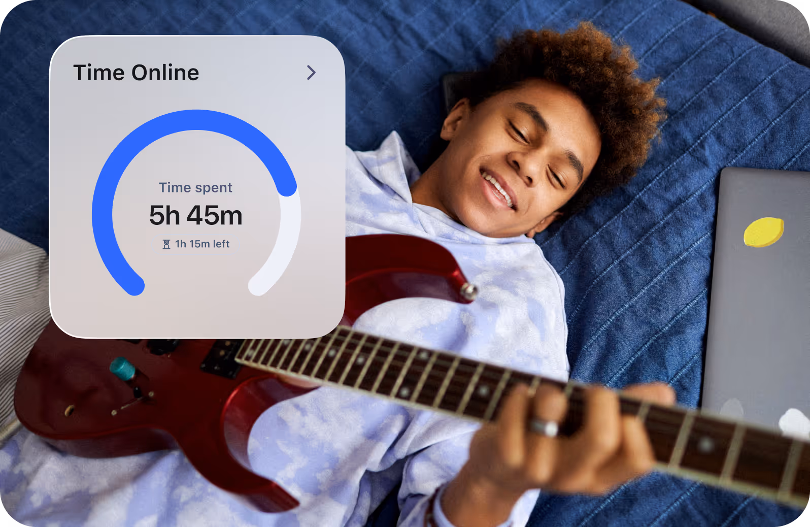 Young teen boy playing an electric guitar laying on bed with laptop closed beside him