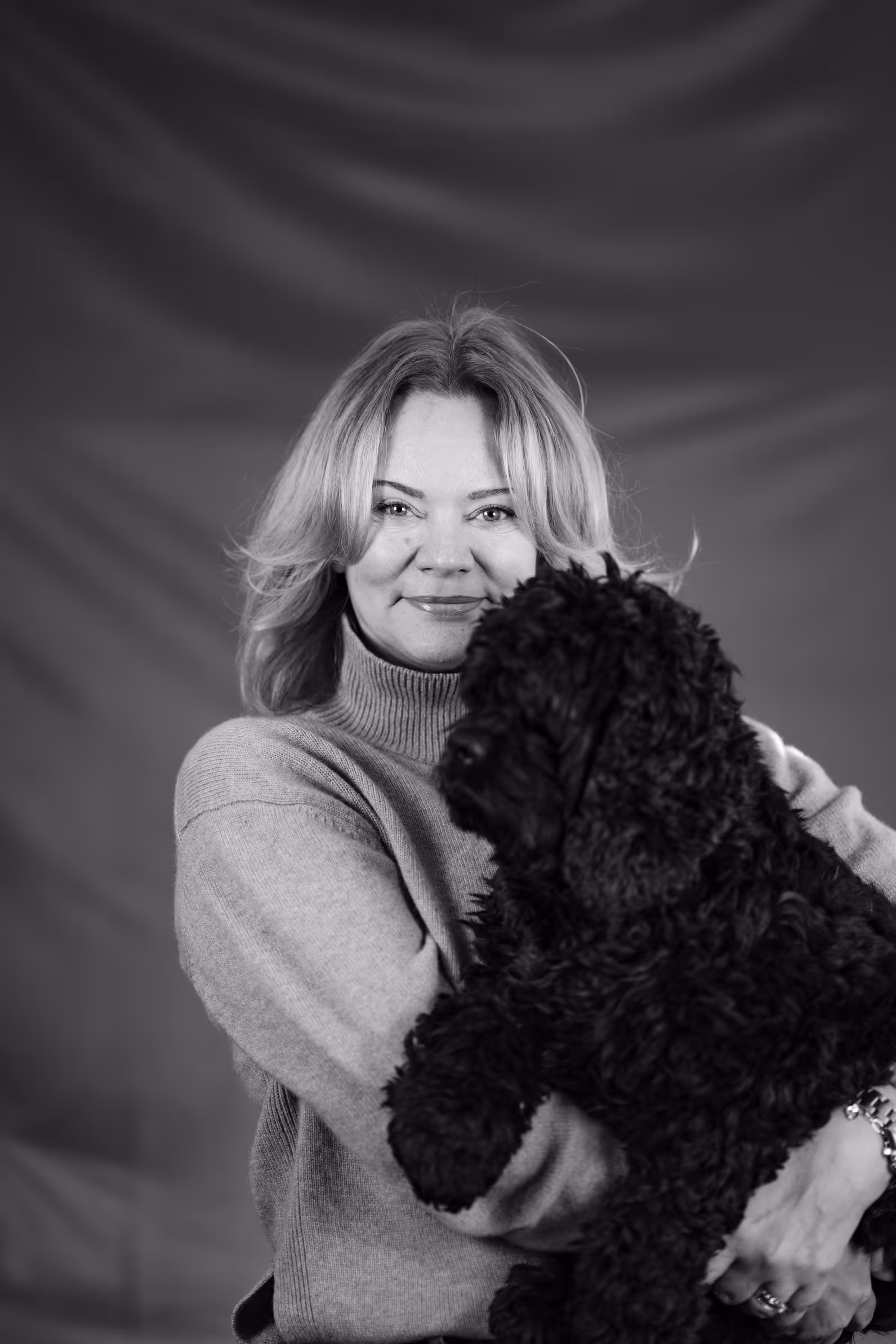 Kvinna i stickad tröja som håller en stor, fluffig svart hund i famnen mot en enfärgad bakgrund.