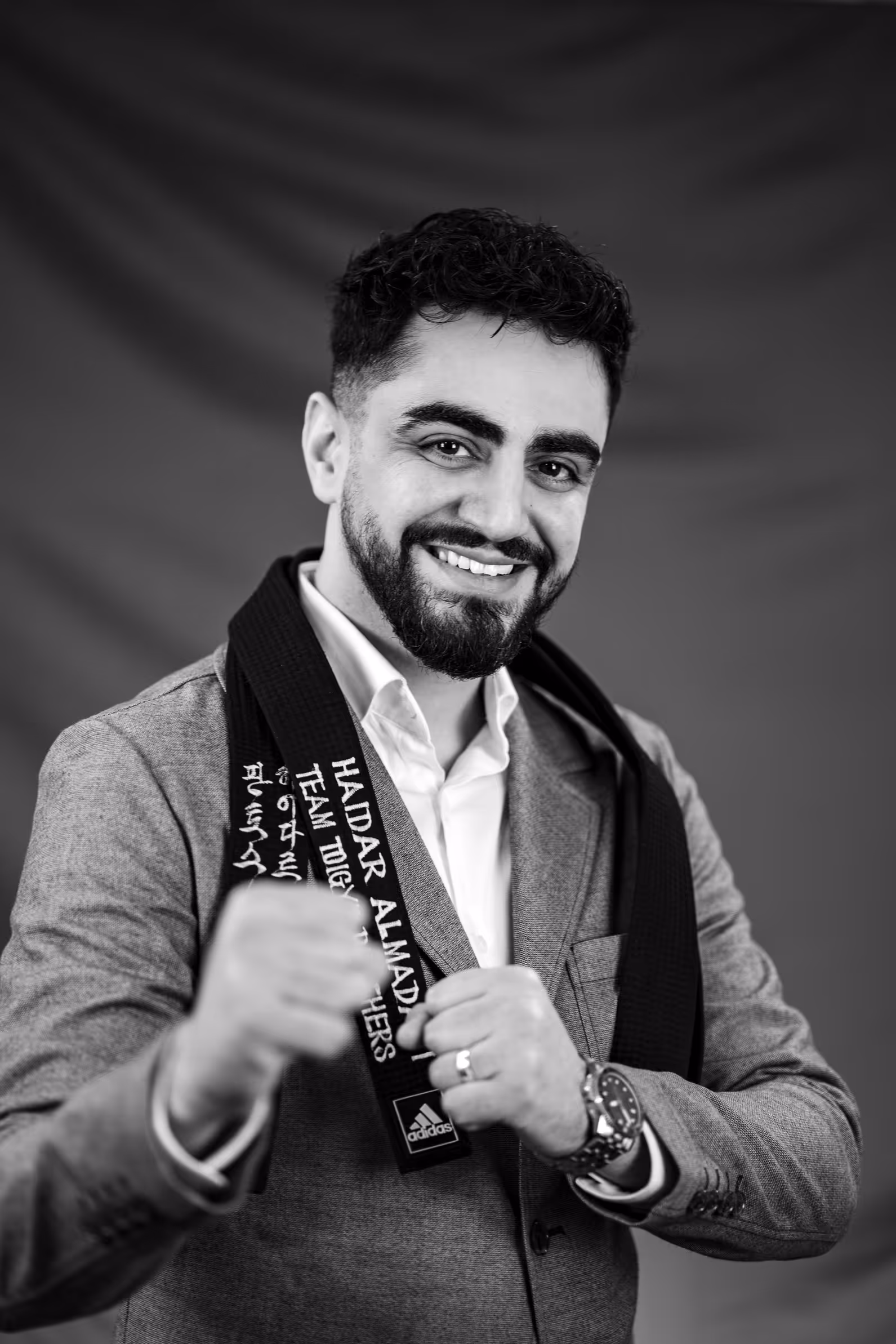 Leende man med skägg i kavaj håller upp ett svart bälte med broderad text och gör en boxningspose.