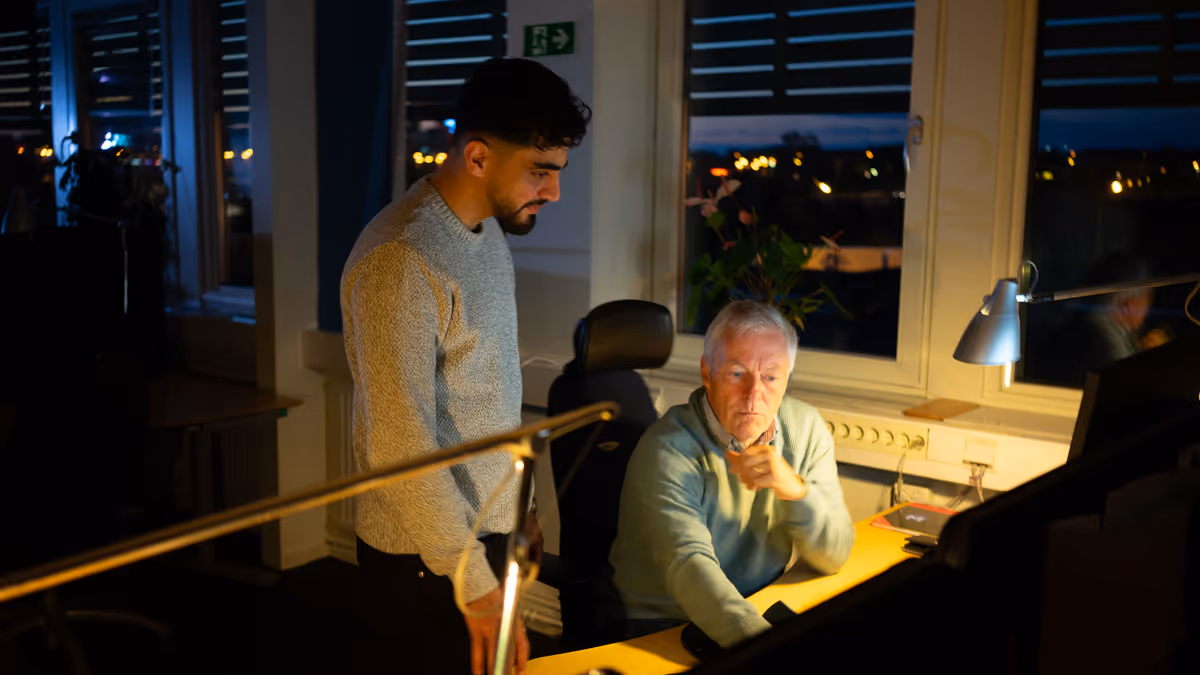 Två män i ett mörkt kontor sent på kvällen, en äldre man sitter vid skrivbordet medan en yngre man står bredvid och tittar på skärmen.