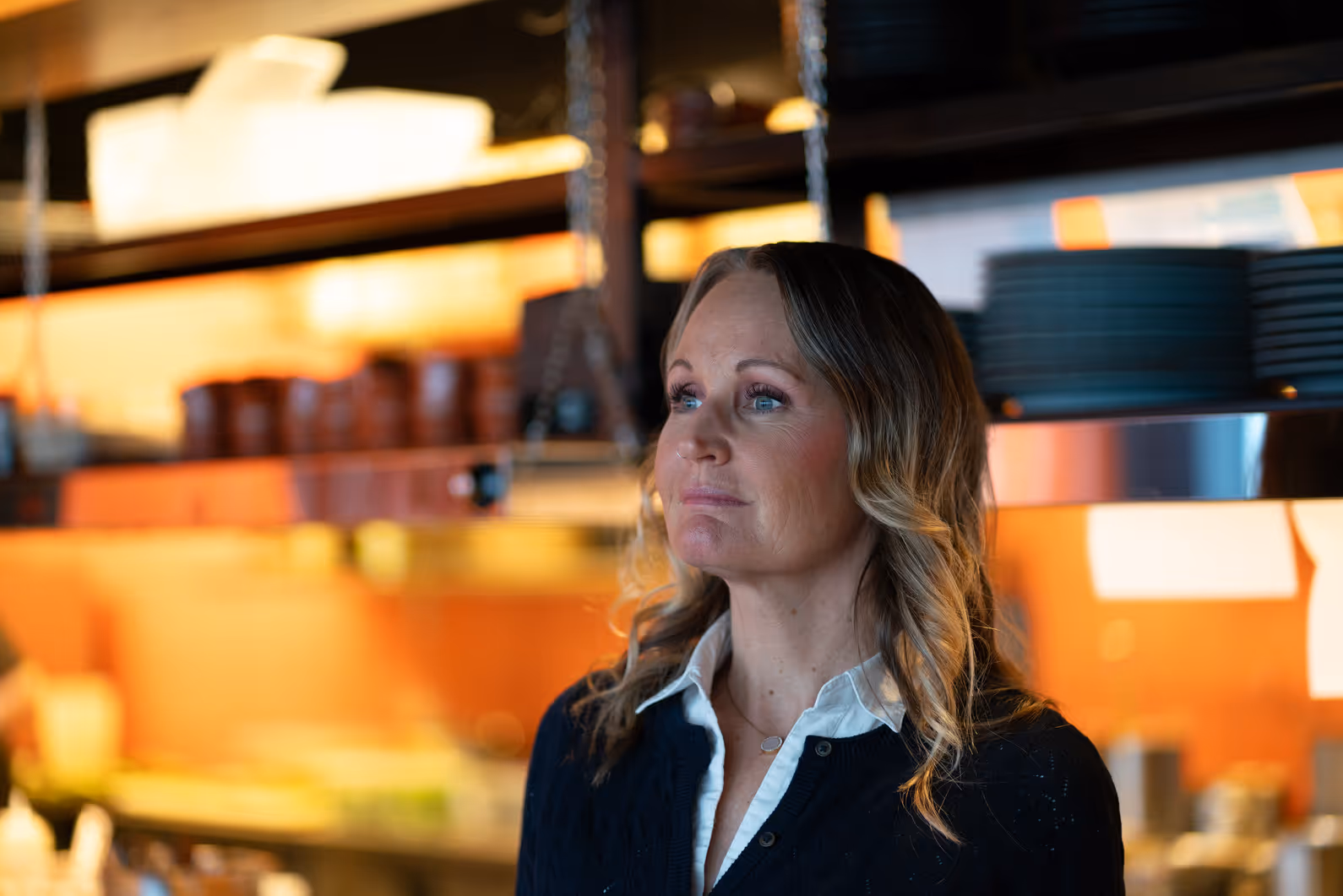 Kvinna med blont lockigt hår och blå tröja som tittar fundersamt mot höger i ett kök.