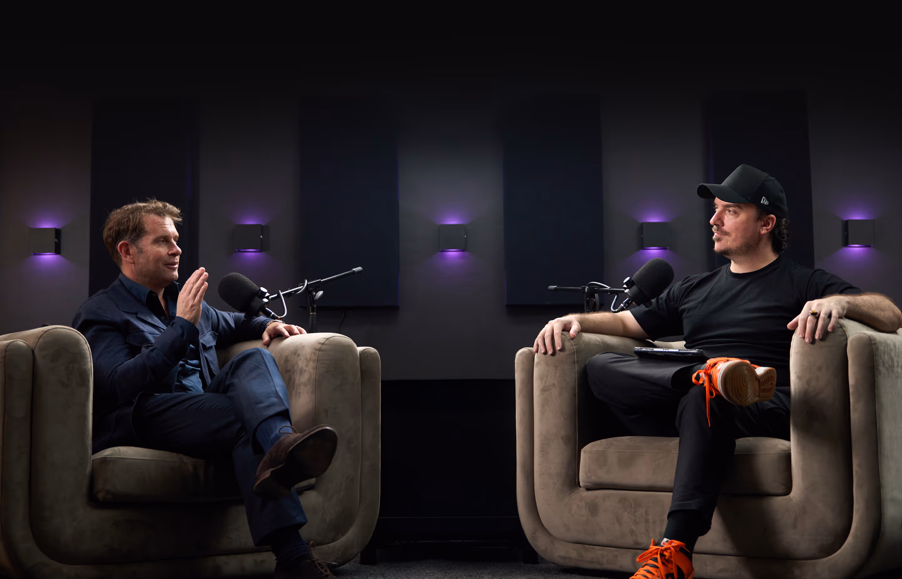 Two men seated in armchairs talking into microphones in a modern podcast studio.
