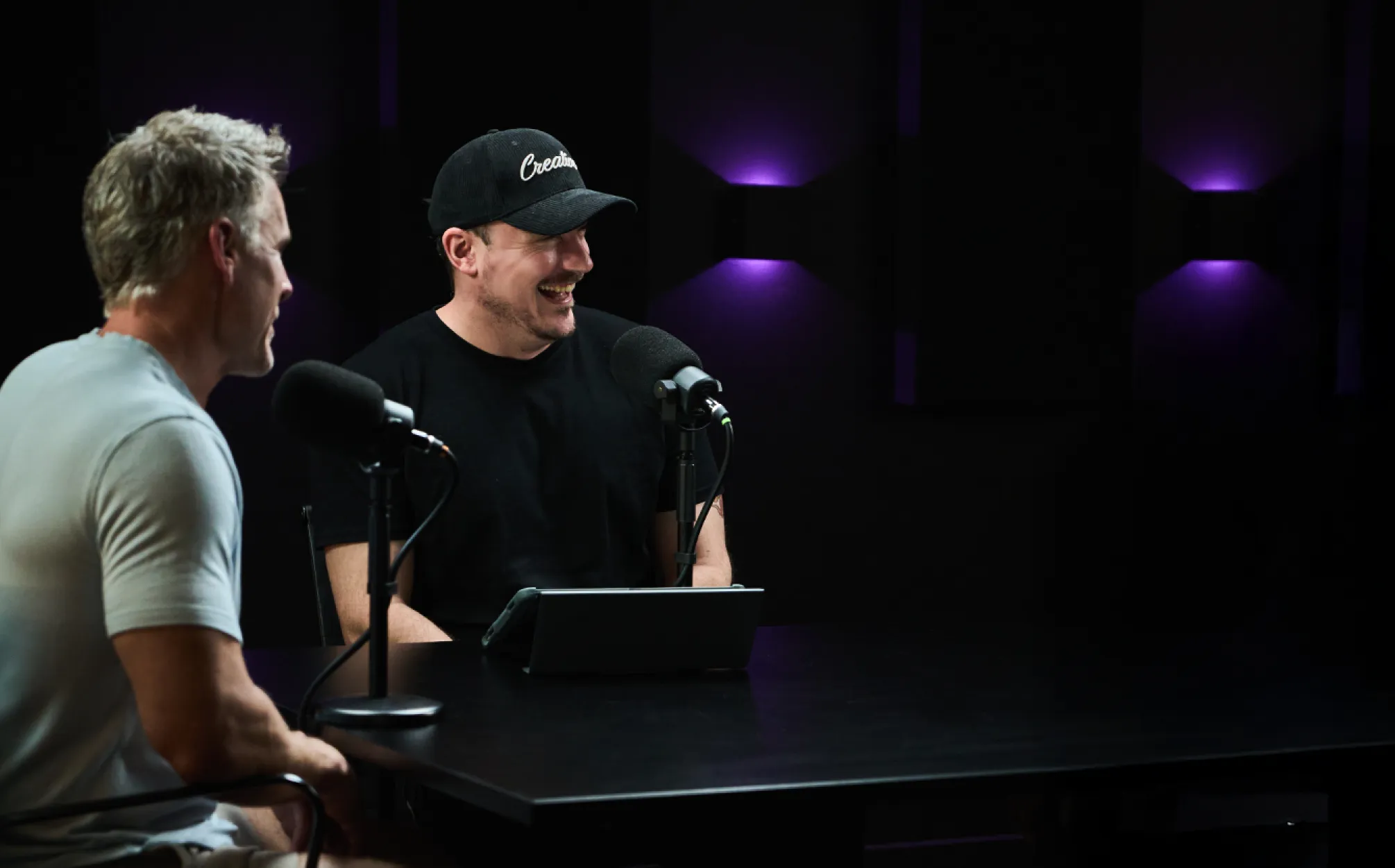 Two men sitting at a table with microphones, recording a podcast in a dimly lit room with purple lighting on the walls.