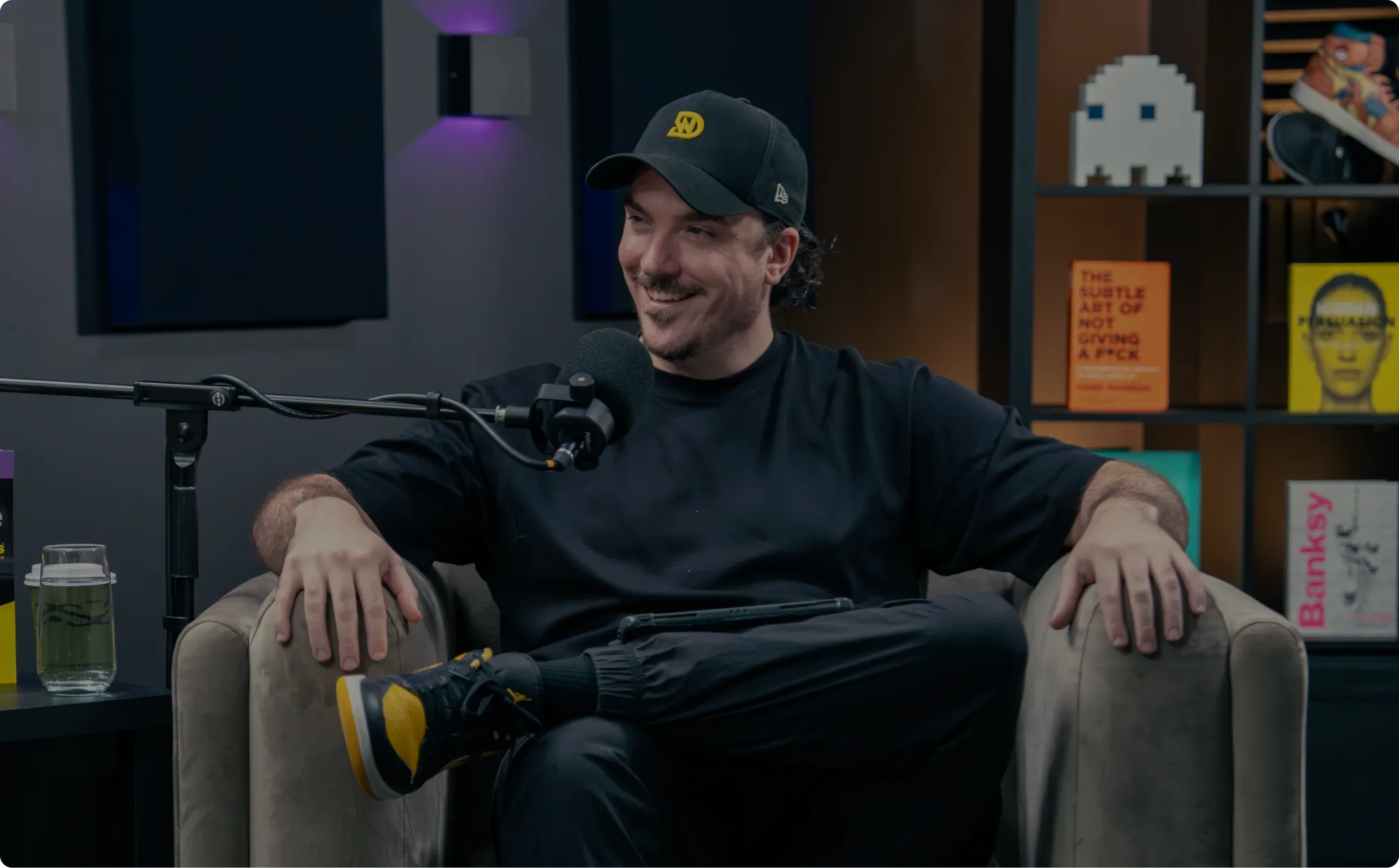 Smiling man in black cap and dark clothes sitting on a chair with a microphone in front.